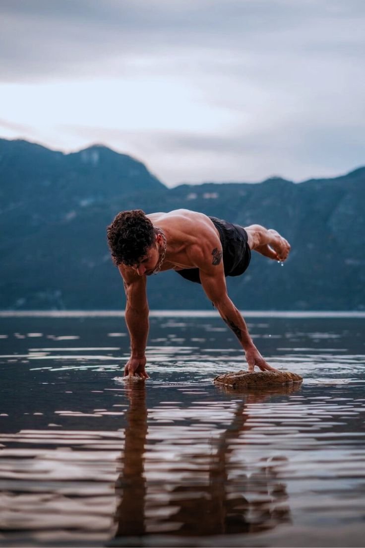 A muscular athlete performs a one-arm plank on a rock in a lake, demonstrating full range of motion and balance.