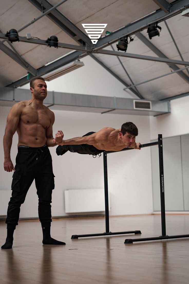 Two men practicing calisthenics in a gym, one holding a static hold on parallel bars while the other observes, highlighting the benefits of daily calisthenics for strength and fitness.