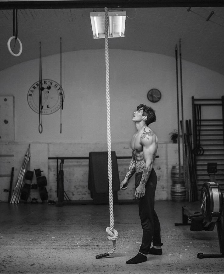 Man standing in a gym, looking up at a rope for climbing, symbolizing dedication and focus in functional fitness training.